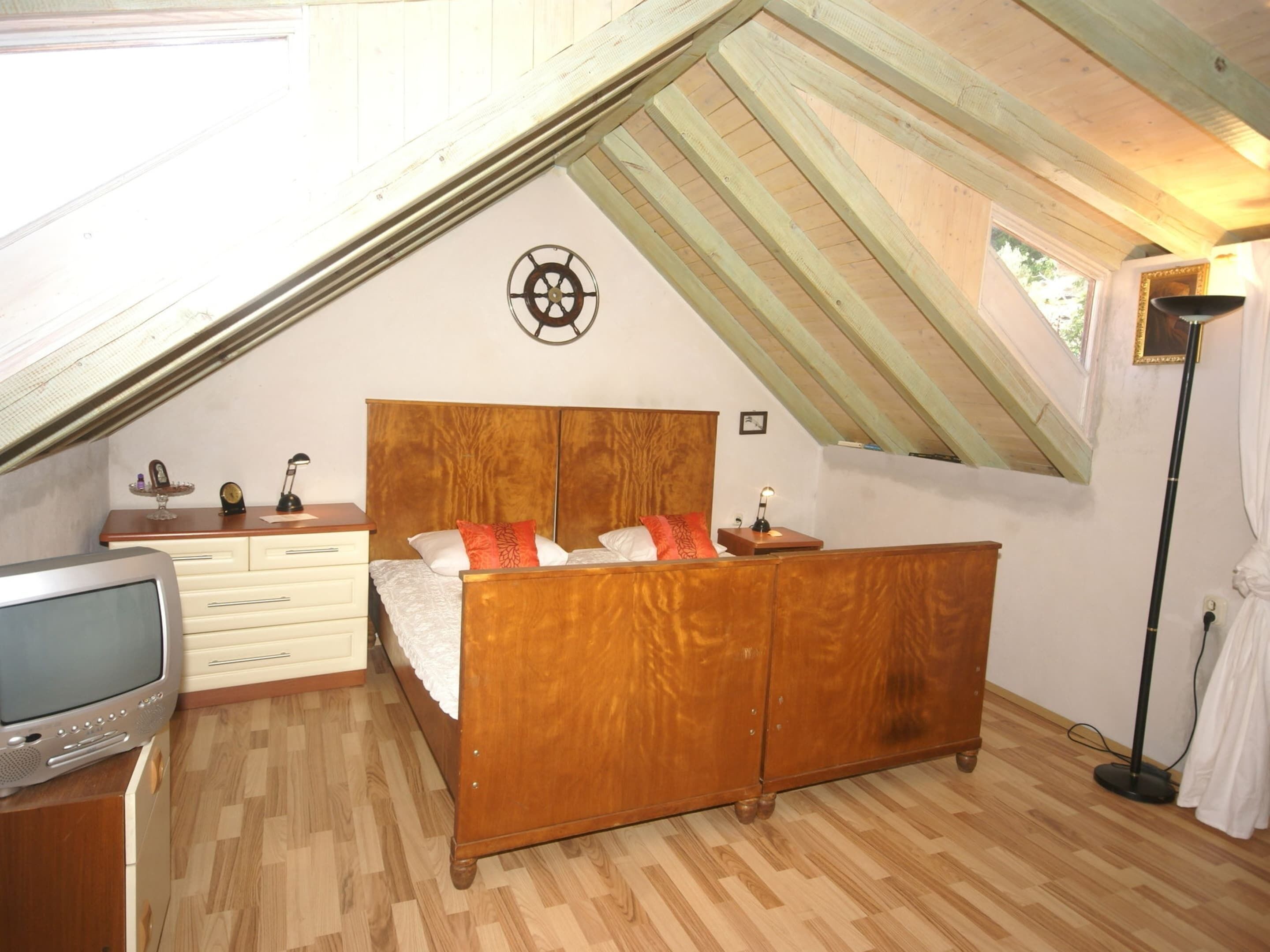 A bedroom with a sloped ceiling, a wooden bed, a dresser, a television, and a small table with a lamp.