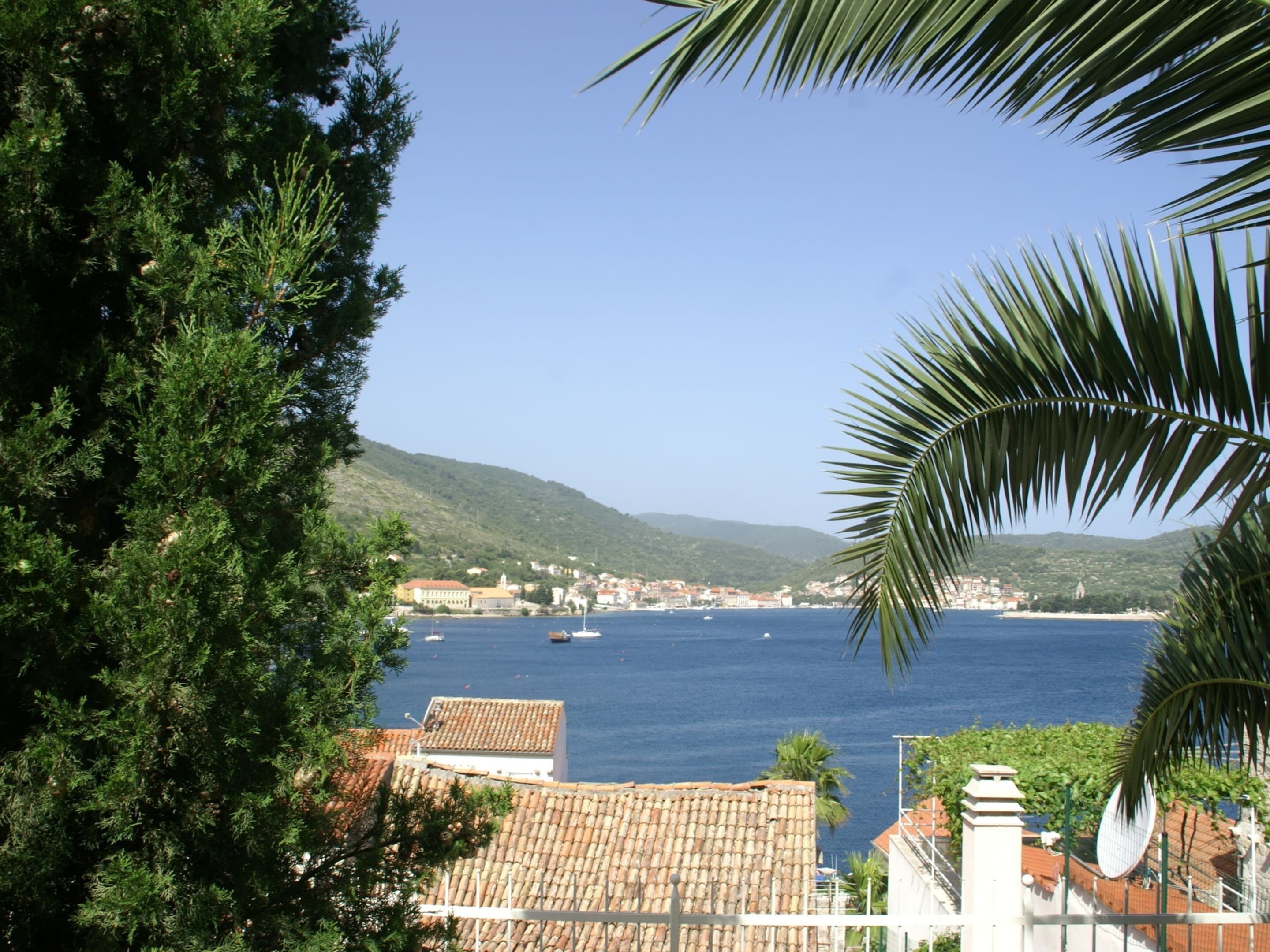 A coastal town with terracotta-roofed buildings, a clear blue sea, and a hilly backdrop.