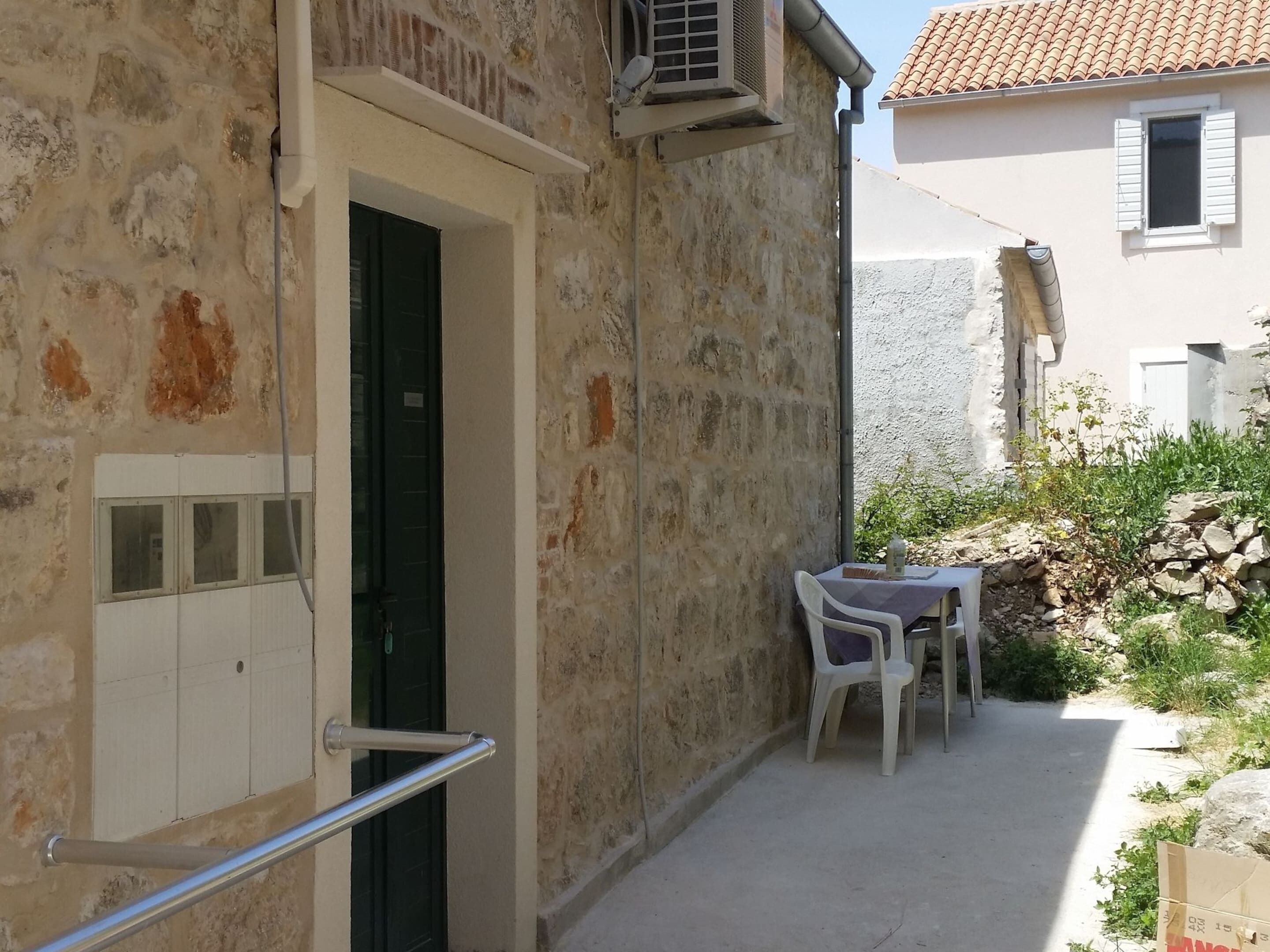 A narrow outdoor corridor with a stone wall, a metal handrail, a small table with a chair, and a window with shutters.