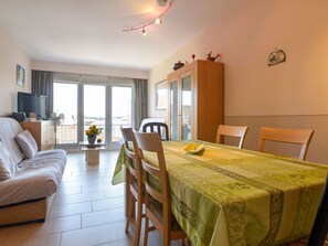 A cozy living room with a sofa, dining table, and chairs. There is a TV, a fan, and a window with curtains. - Simplistic Apartment in Blankenberghe near Belgium Pier (Blankenberge)