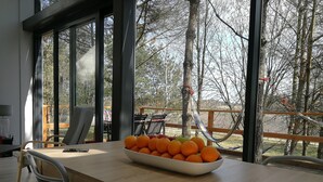 A modern dining area with a wooden table, a bowl of oranges, and a view of a deck and trees. - Glass Cottage in Jelenska Huta With Terrace (Szemud)