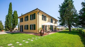A two-story yellow building with green shutters, a patio with outdoor seating, and a well-maintained lawn. - Belvilla by OYO San Francesco Vermentino (Lajatico)