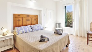 Un dormitorio con cabecera de madera, una cama con ropa de cama floral, una mesita de noche con lámpara y vistas al exterior a través de una ventana.