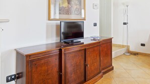 A wooden cabinet with a flat-screen TV on top, a framed picture above it, and a floor lamp to the right. - Belvilla by OYO Vega (Caprino Veronese)