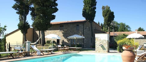 Una piscina con sillones de descanso y sombrillas, un edificio de piedra con tejado de baldosas y altos árboles.
