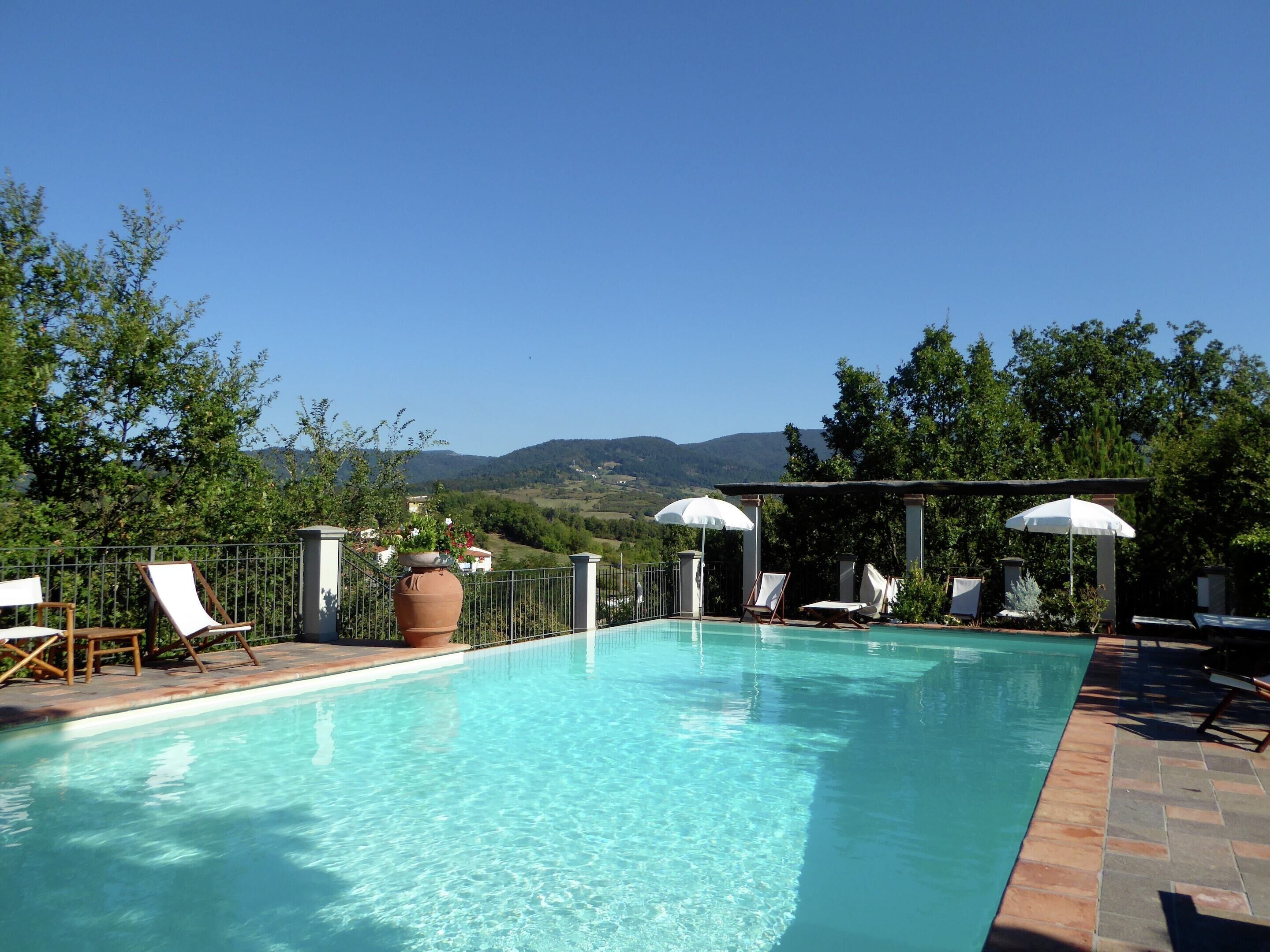 A clear swimming pool with lounge chairs and umbrellas, surrounded by greenery and a view of distant hills.