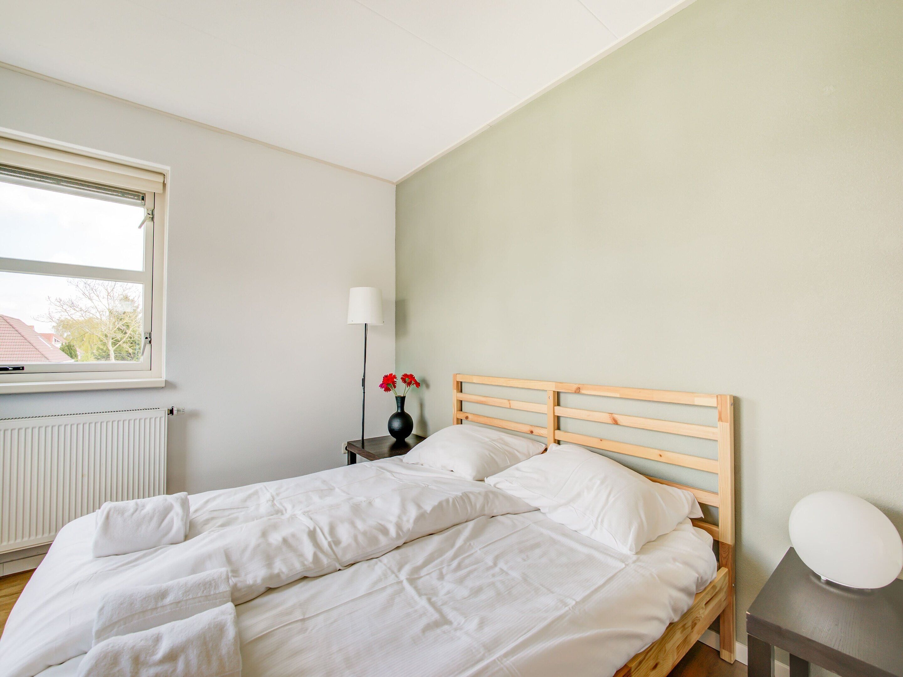 A bedroom with a wooden bed, white bedding, a nightstand with a lamp and vase, and a window with blinds.