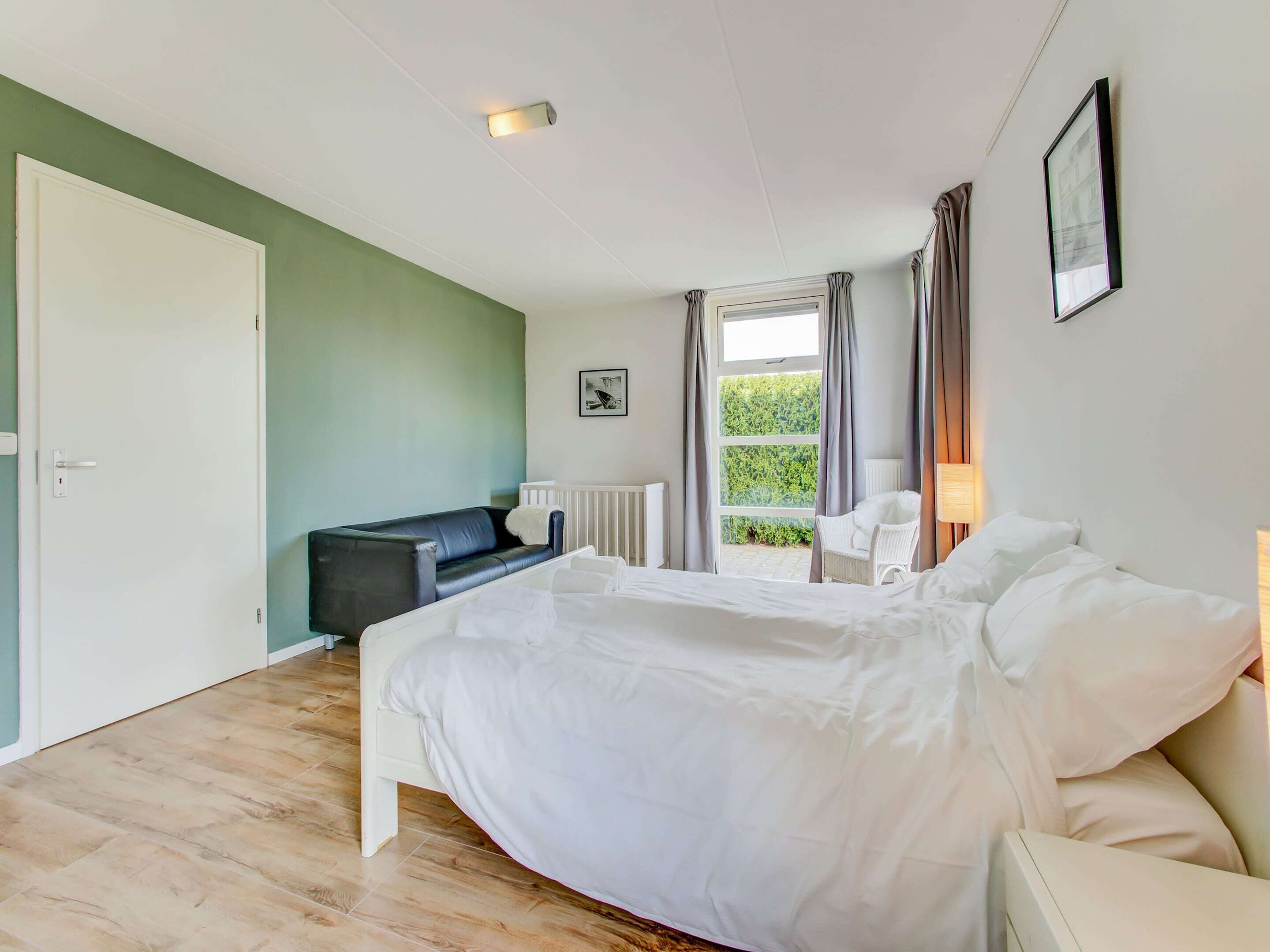Een slaapkamer met een bed, een witte stoel, een zwarte bank, een raam met gordijnen en een radiator.