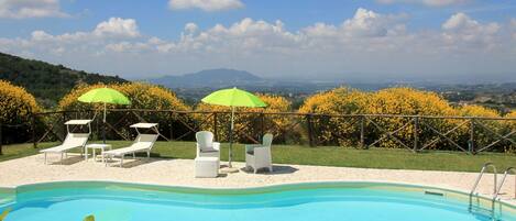 A pool area with lounge chairs and umbrellas, surrounded by a fence and a view of the landscape.