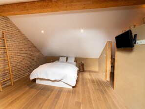 A bedroom with a sloped ceiling, wooden floor, a bed with white bedding, a television on the wall, and a brick wall. - Bertrix Charming 2-bedroom Holiday Home (Bertrix)