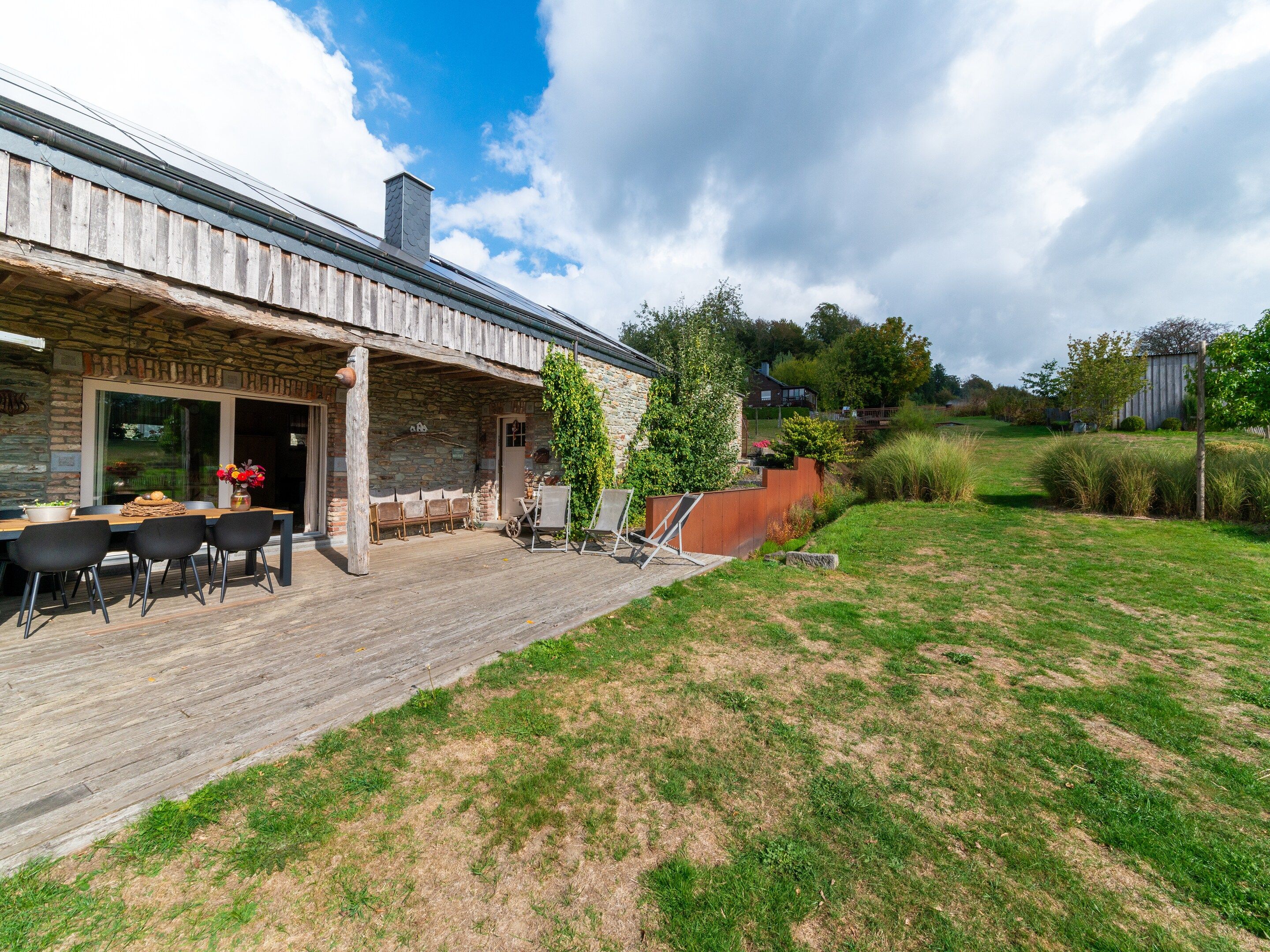 A rustic stone building with a wooden deck, outdoor seating, and a grassy yard.