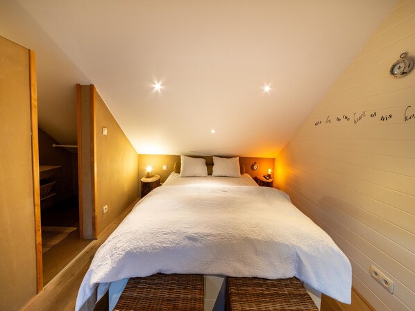 A bedroom with a large bed, a bedside table with a lamp, a wall clock, and a staircase leading to a closet.