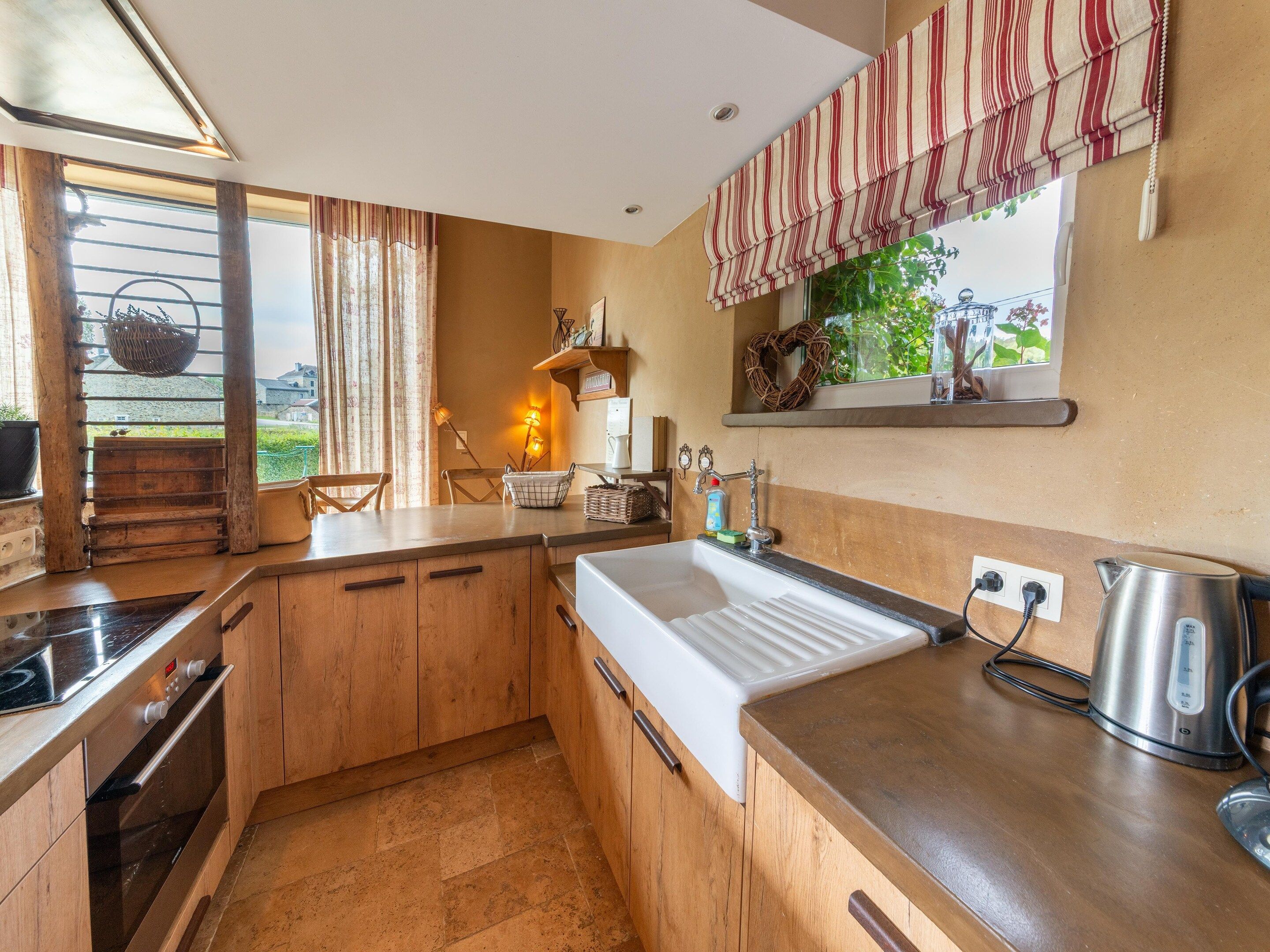 A kitchen with wooden cabinets, a white sink, a stainless steel kettle, and a window with a view of greenery.