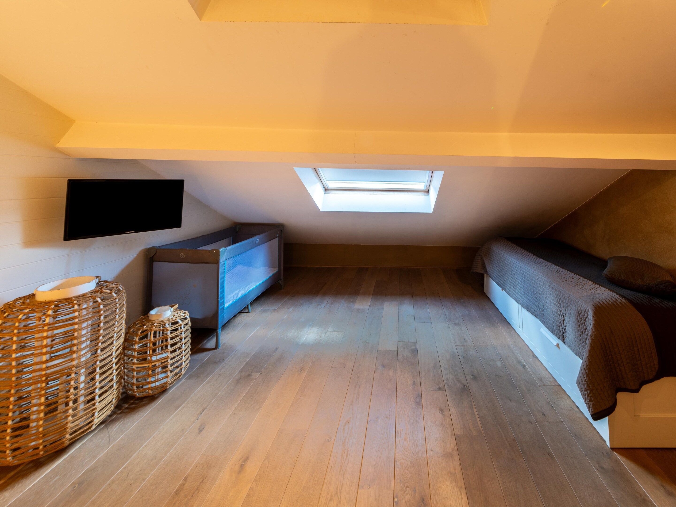 Ein kleines Zimmer im Dachgeschoss mit schrägen Wänden, einem an der Wand befestigten Fernseher, einem Etagenbett und zwei Korbwannen auf dem Boden.