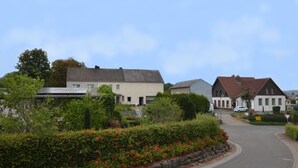 A residential street with houses, a winding road, and a hedge with red flowers. - Ruhlen: Schneifel Escape (Auw bei Prüm)