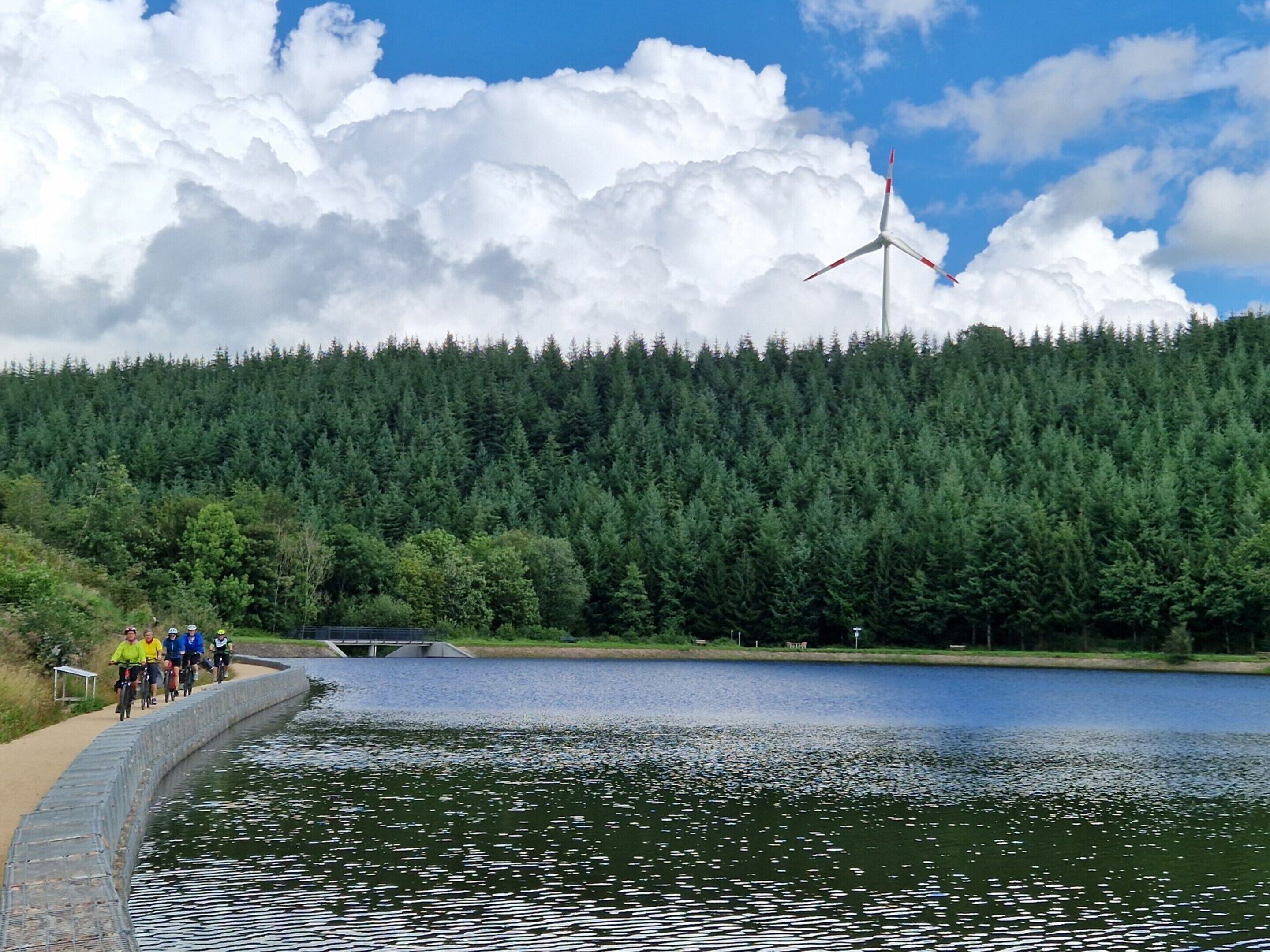 A lake with a forested hill, a wind turbine, and a paved path with cyclists.