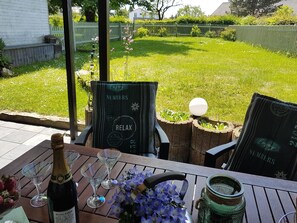 Um pátio com uma mesa posta para dois, apresentando uma garrafa de champanhe Duval-Leroy, duas taças de vinho, um vaso de flores e uma jarra de cerâmica verde.