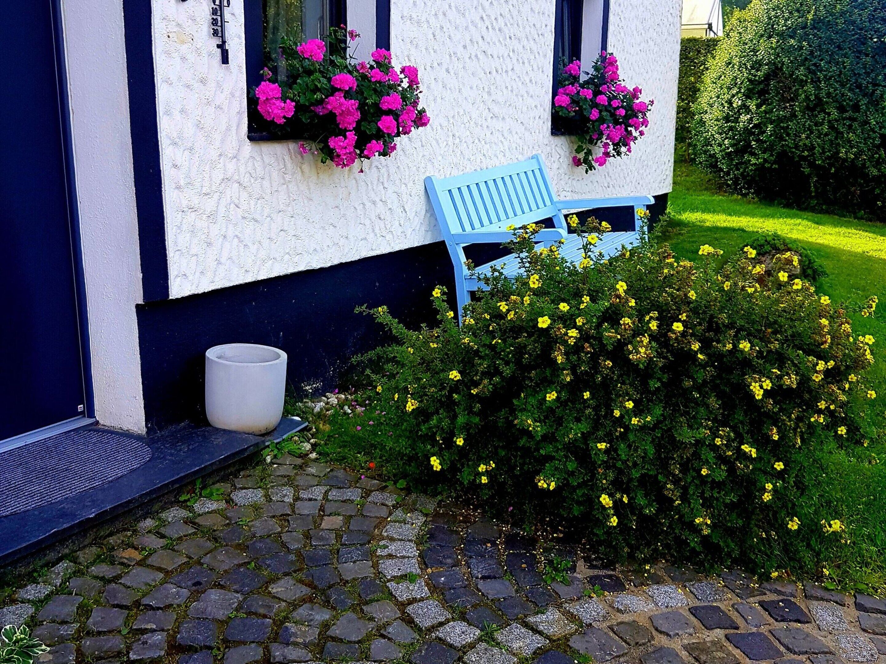 Un edificio con fachada blanca y detalles oscuros, un banco azul en el exterior y una jardinera con flores rosas.