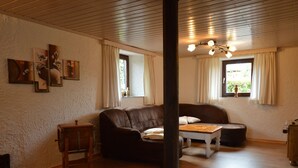 A living room with a brown leather sofa, a wooden coffee table, and a wooden chair. There are two windows with curtains, a wooden ceiling, and a wall with framed artwork. - Ruhlen: Schneifel Escape (Auw bei Prüm)