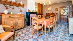 Een eetgedeelte met een tafel voor zes personen, omringd door houten stoelen. Achterin is een open haard te zien, en er hangen diverse keukengerei aan de muur.