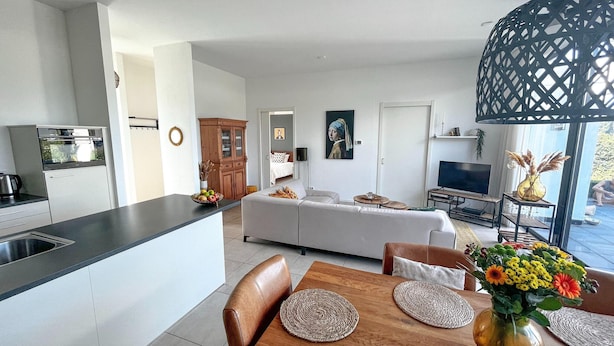 Eine moderne Küche mit Kochinsel, ein Essbereich mit Holztisch und Stühlen, ein Wohnzimmer mit Sofa und Fernseher sowie ein großes Fenster für natürliches Licht.
