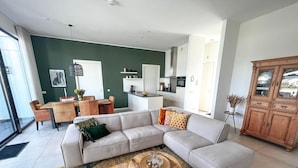 Ein modernes Wohnzimmer mit einem Ecksofa, einem Essbereich mit Tisch und Stühlen, einer Küche im Hintergrund und einer großen Holzkommode.