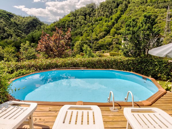 An outdoor swimming pool with wooden deck and lounge chairs, surrounded by lush greenery and hills.