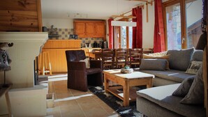 A cozy living room with a sofa, chairs, a wooden coffee table, and a kitchen area with wooden cabinets and a window with red curtains. - Chalet Chocolat chaud 4 rooms - 90m², sleeps 7 in the heart of Belleville (st martin de Belleville)
