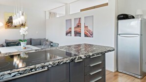 A modern kitchen with a granite countertop, dark cabinetry, and a built-in oven. There are three framed pictures on the wall, a refrigerator, and a dining area with a gray sofa and a vase of white flowers. - Apartment Innsbruck Near Bergisel Ski Jump (Innsbruck)