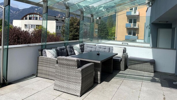 A covered outdoor seating area with wicker furniture and a glass roof, overlooking a mountain and residential buildings. - Apartment Innsbruck Near Bergisel Ski Jump (Innsbruck)