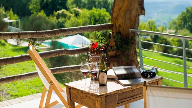 A wooden table with wine glasses, a bottle, and a book on a balcony overlooking a lush landscape.