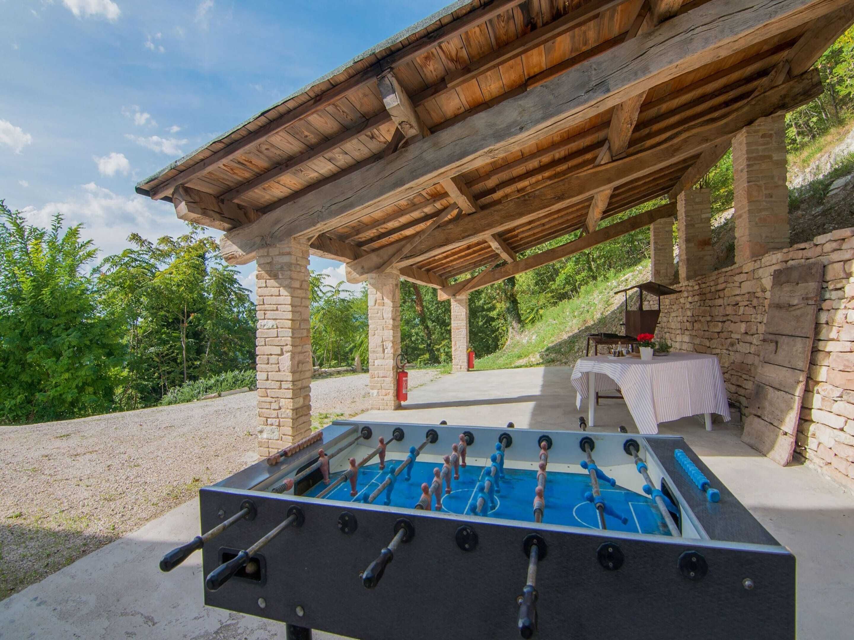 An outdoor foosball table under a wooden pergola with stone pillars, set against a backdrop of greenery and a clear sky.