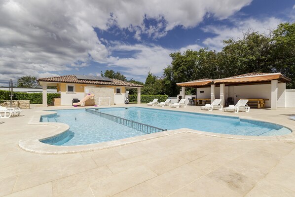 A swimming pool with a tiled deck, lounge chairs, and a covered area with a tiled roof.