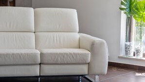 Um sofá de couro branco com base de cromo, posicionado em um quarto com piso de azulejos e uma janela com uma planta.