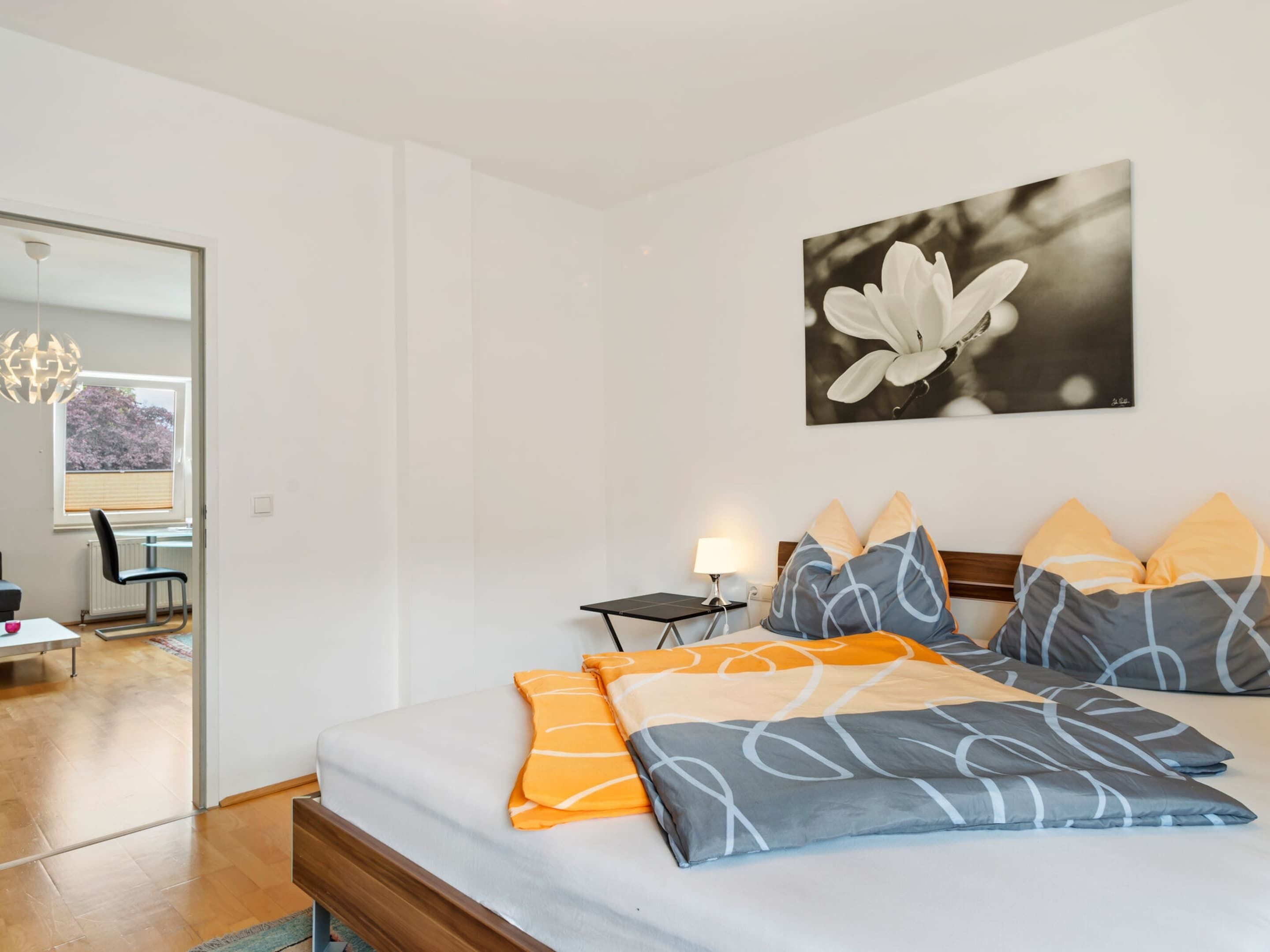 Un dormitorio con cama, almohadas, mesita de noche, lámpara y un cuadro colgado en la pared.