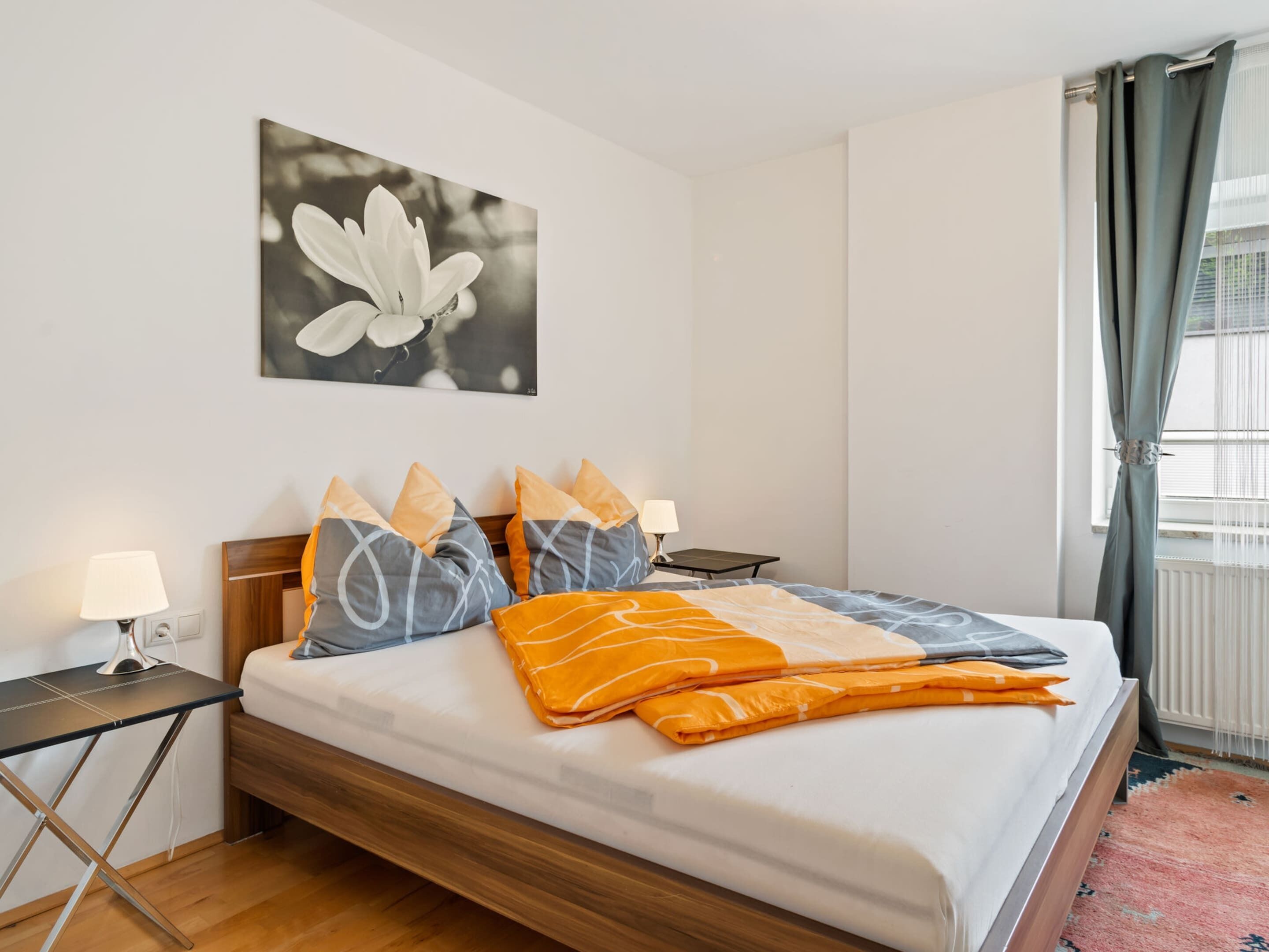 A neatly made bed with a white mattress, grey and orange pillows, and a grey and orange blanket. There is a small bedside table with a lamp, a wall-mounted picture of a flower, and a window with curtains.