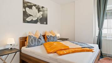 A neatly made bed with a white mattress, grey and orange pillows, and a grey and orange blanket. There is a small bedside table with a lamp, a wall-mounted picture of a flower, and a window with curtains.