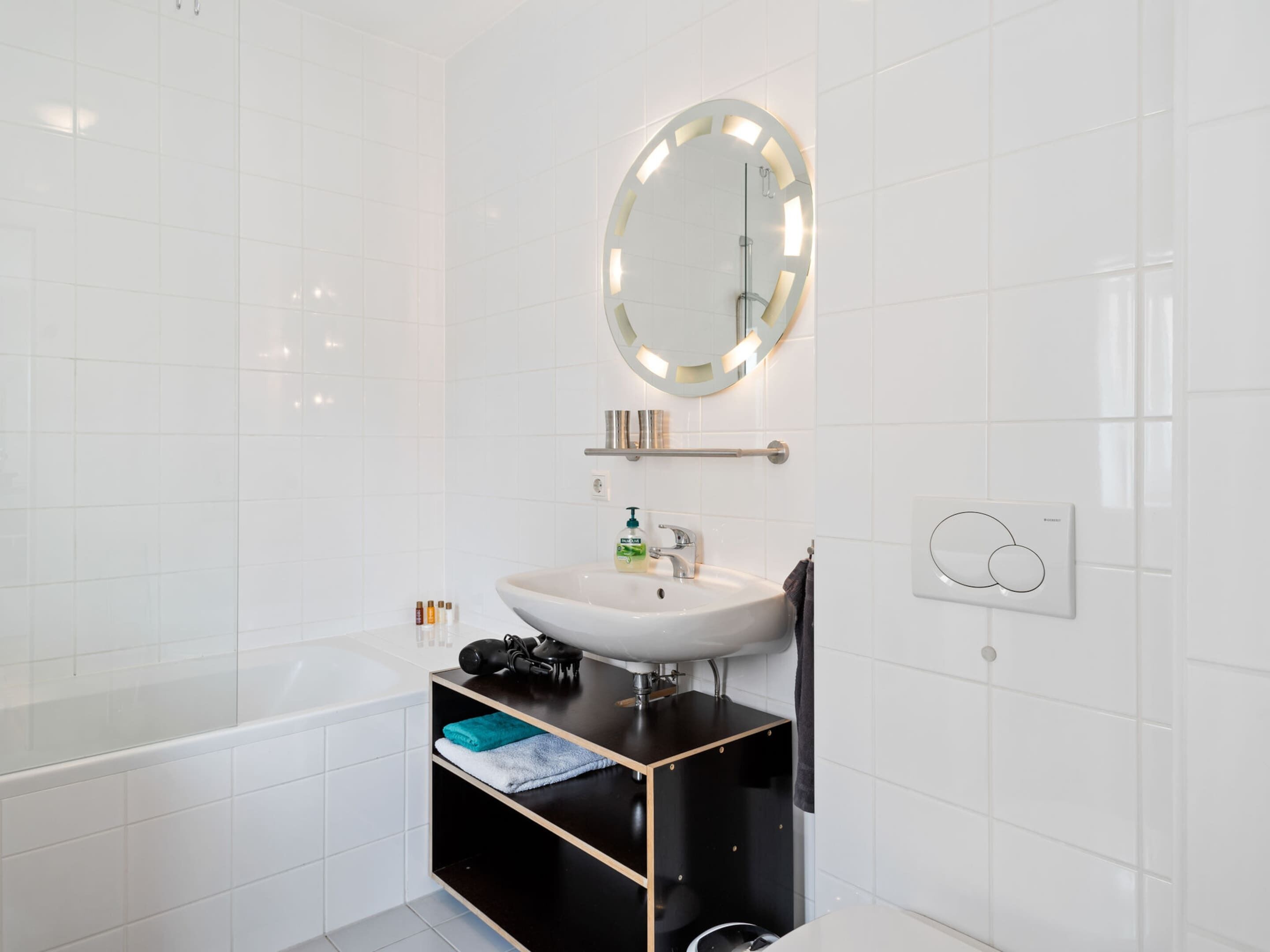 A modern bathroom with a white sink, a mirror with lights, a towel rack, and a black storage unit.