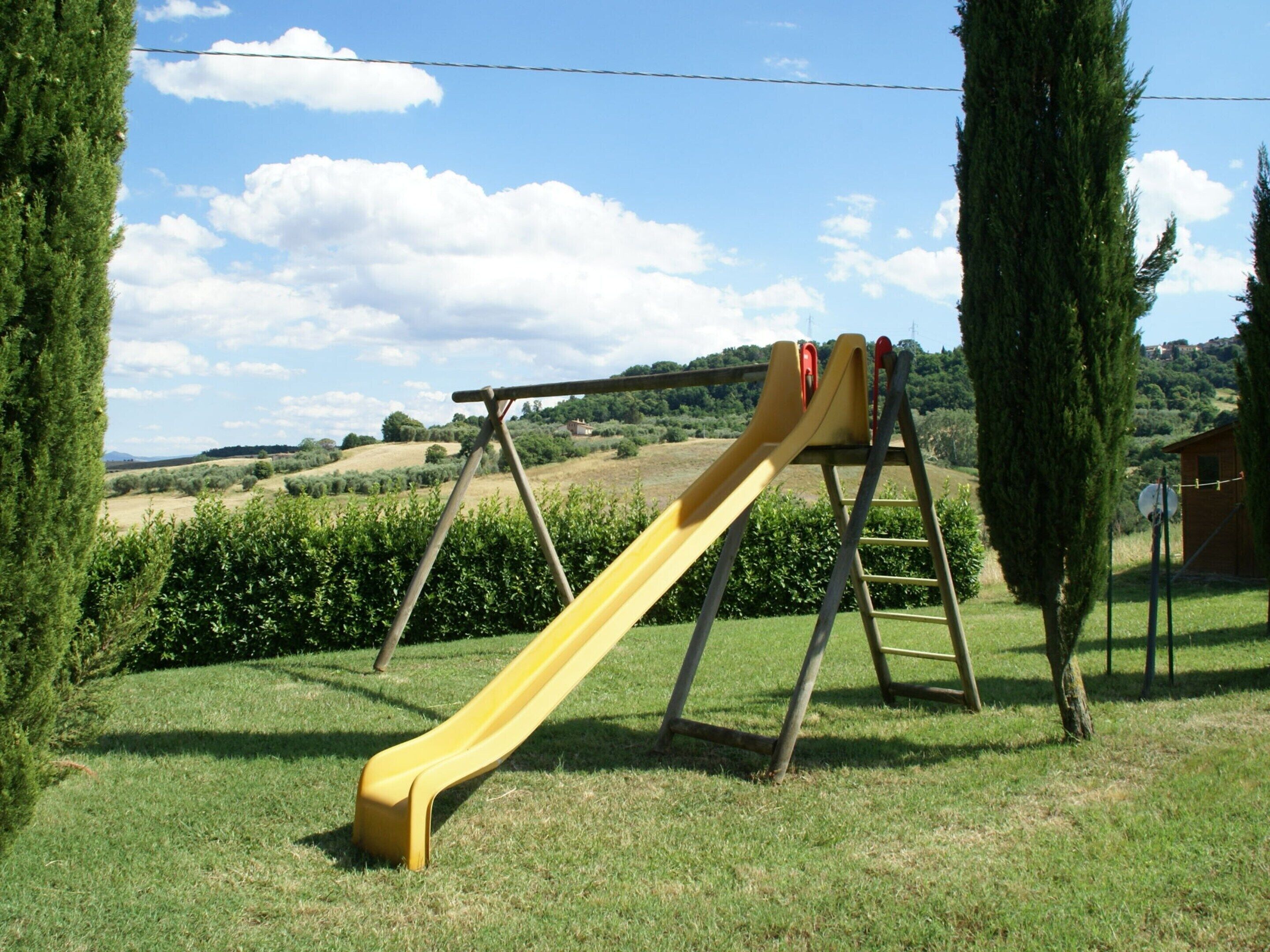 Un parque infantil con un tobogán amarillo y una escalera de madera, rodeado de césped y árboles.