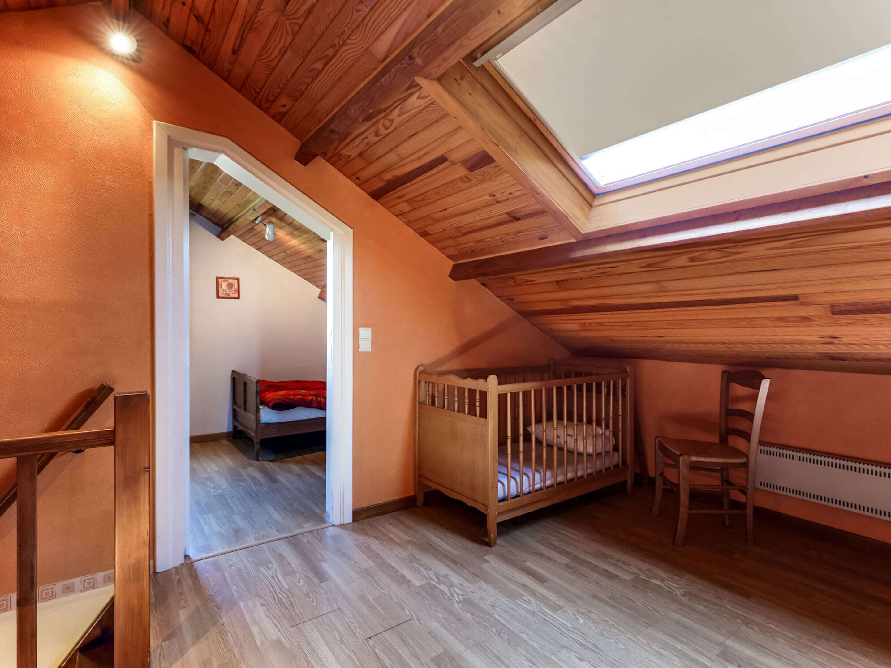 Un dormitorio acogedor en el ático, con techo inclinado, vigas de madera, una cuna pequeña y una silla.