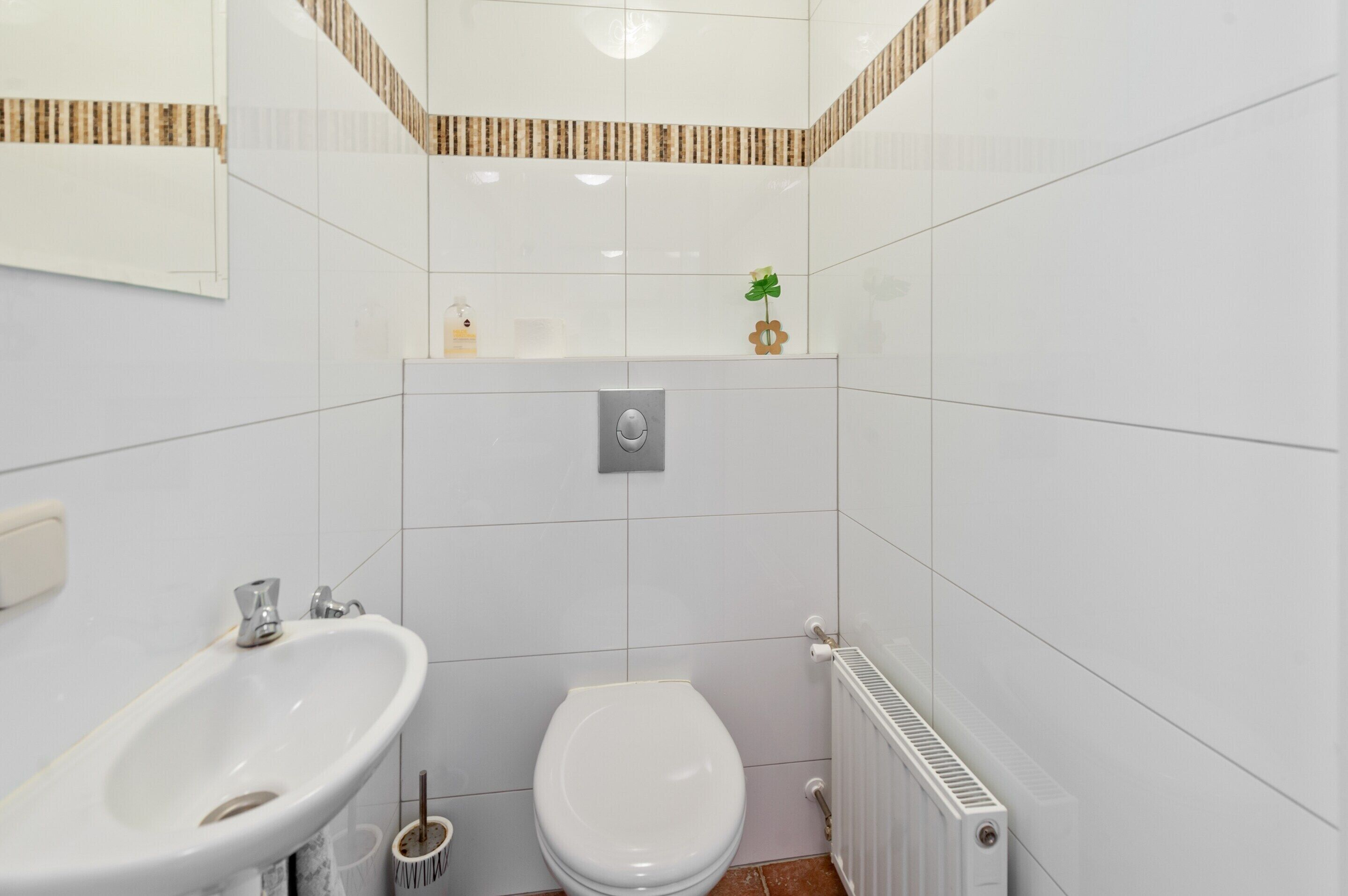 A small, modern bathroom with white tiles, a toilet, a sink, and a radiator.