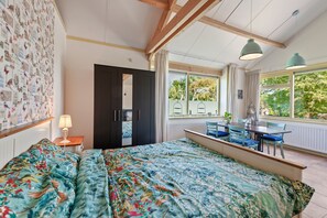Quarto com cama, mesa de jantar com cadeiras, janela com vista para área verde e parede com papel de parede estampado.