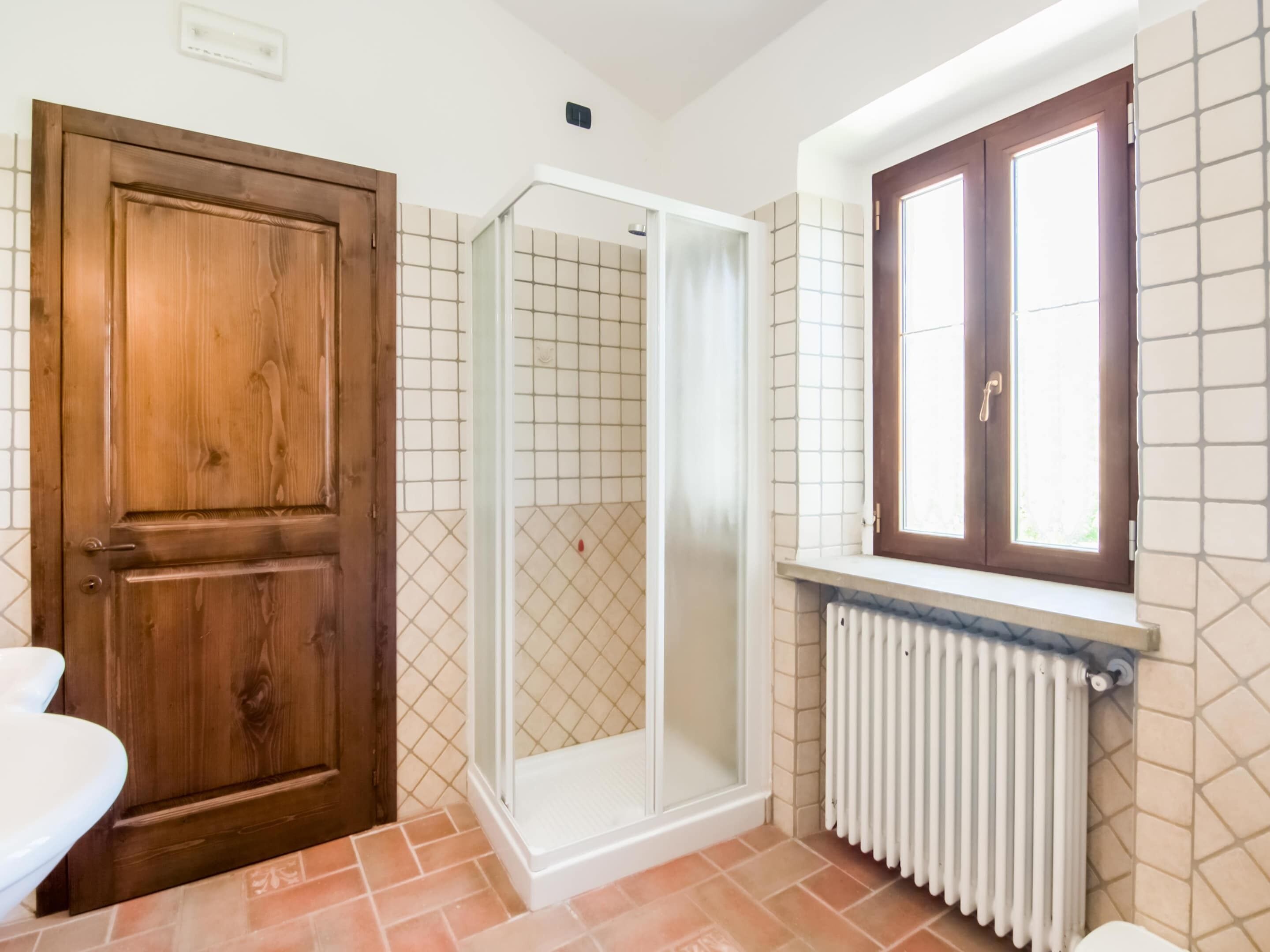 A bathroom with a wooden door, a shower enclosure, a window with a wooden frame, and a radiator.