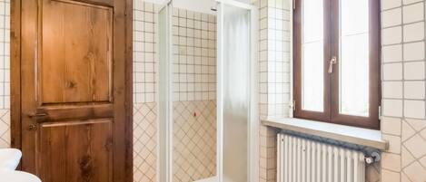 A bathroom with a wooden door, a shower enclosure, a window with a wooden frame, and a radiator.