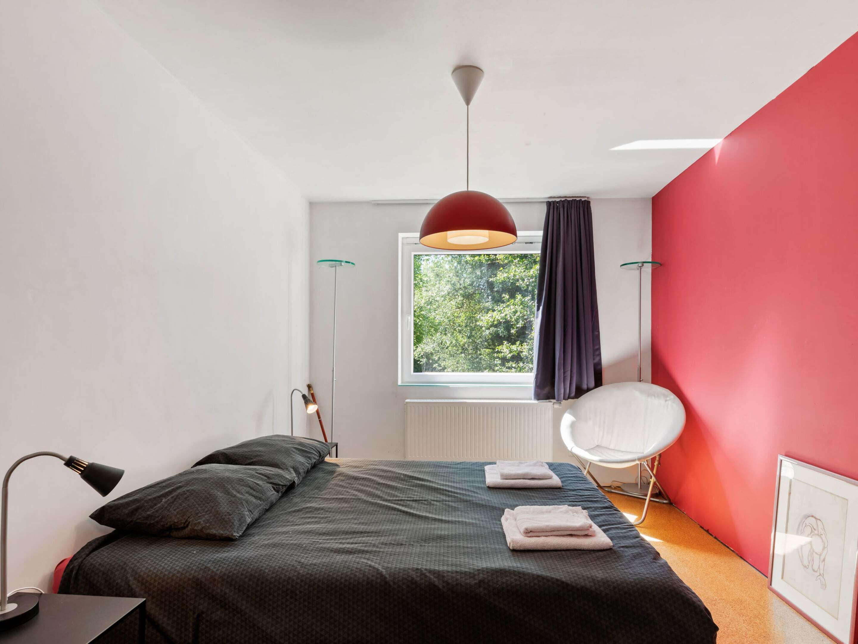 Un dormitorio moderno con cama, lámparas de noche, una ventana con cortinas, una pared de acento roja y una silla.