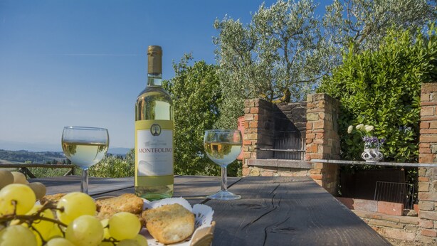 A table with a bottle of Monteolivo wine, two wine glasses, a basket of grapes, and a biscuit. - Belvilla by OYO Fienile (Castelfiorentino)