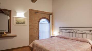 A bedroom with a brick wall, a bed with a metal headboard, a window with curtains, a wooden ceiling, and a framed picture on the wall.
