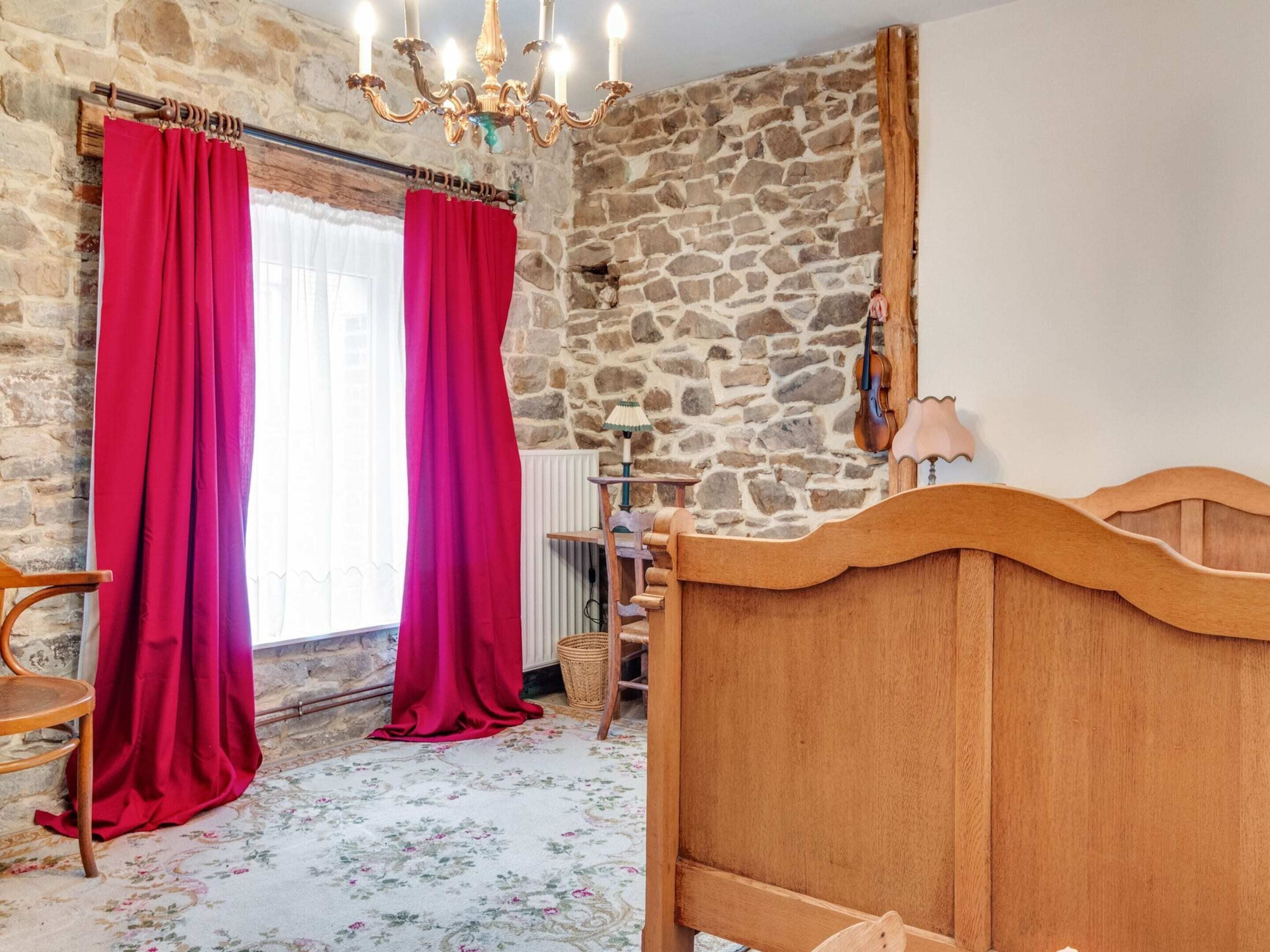 Un dormitorio con pared de piedra, cama de madera, cortinas rojas, candelabro y radiador.