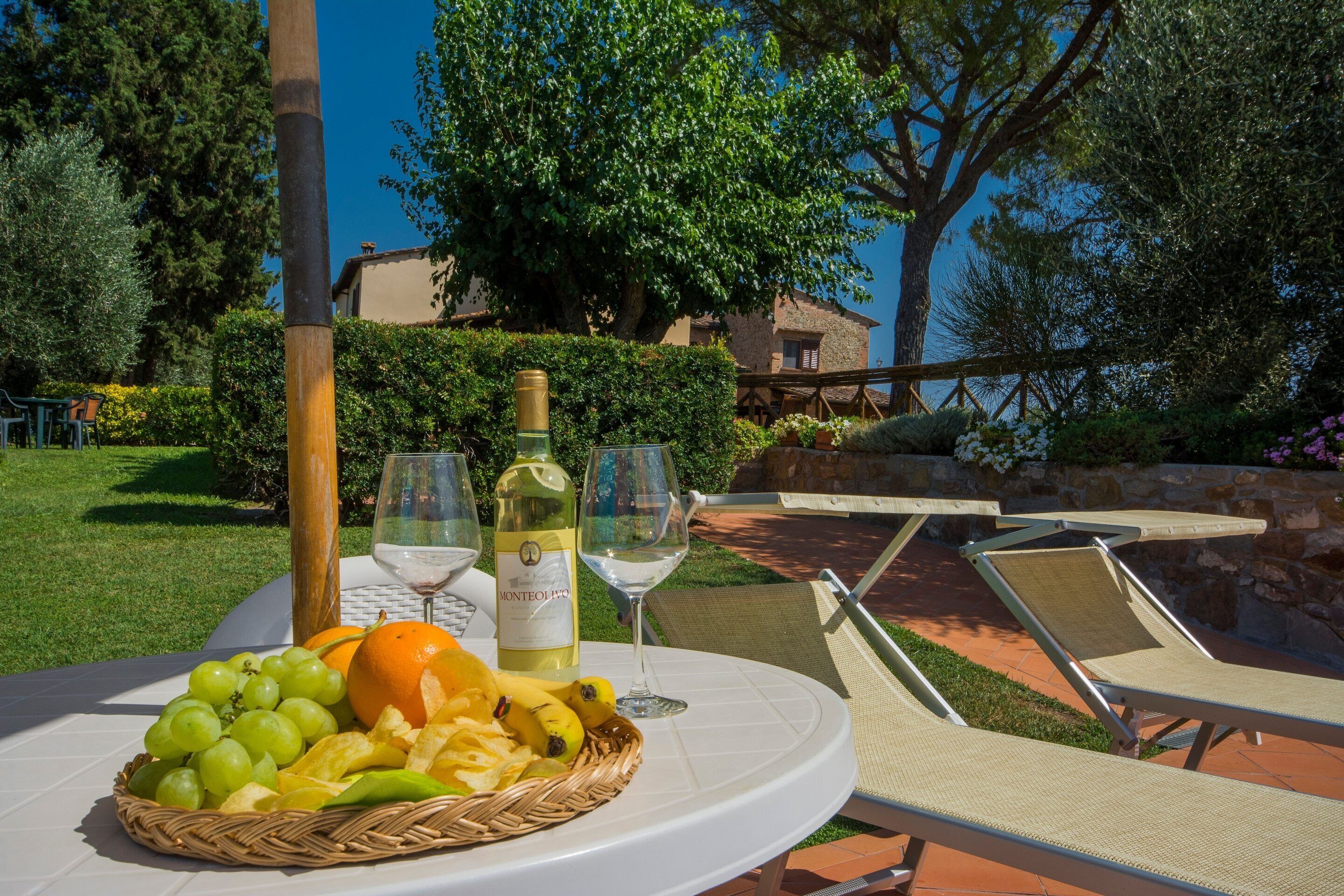 A table with a basket of fruits, a bottle of wine, and glasses, set outdoors with lounge chairs and a view of a building and trees.
