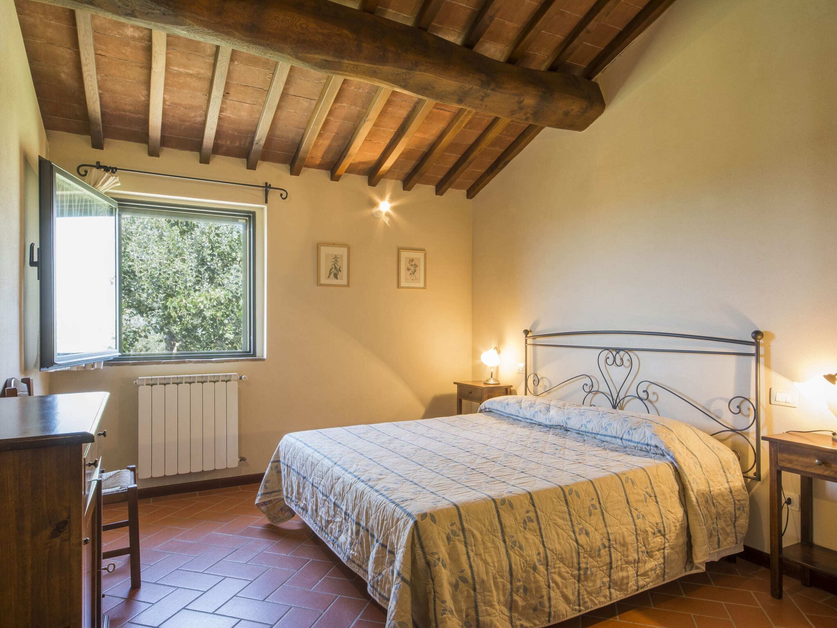 A bedroom with a wooden ceiling, a bed with a metal frame, a window with a view of trees, a radiator, and a small bedside table with a lamp.